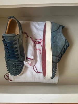 Blåa Louboutin sneakers med nitar - Unika blåa sneakers från Christian Louboutin i mocka med coola nitar på tån och hälen. Skorna har klassisk röd sula, vita kanter och mörkblå snören. Perfekta för dig som vill sticka ut med en edgy och lyxig stil.