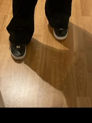 Svarta sneakers med gulddetalj - Stilrena svarta sneakers med vita sulor och guldfärgade detaljer på snörningen. Skorna har en klassisk låg profil och är tillverkade i slätt material som ger en clean look. Perfekta för dig som gillar enkel men snygg design.