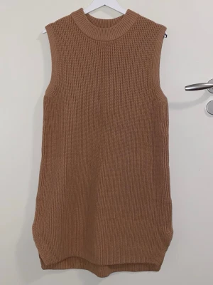 Beige stickad ärmlös klänning - Säljer en beige stickad ärmlös klänning med hög ribbad krage. Klänningen har rak passform och är gjord i ett mjukt, grovstickat material. Perfekt att styla med en skjorta under eller över ett par tights.