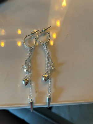 Silvriga örhängen med hjärta och kedjor - Säljer ett par silvriga örhängen med små ringar, hängande kedjor, hjärtformade detaljer och spetsiga hängen. Örhängena har en modern och edgy look med flera tunna kedjor som ger extra rörelse. Perfekt för dig som gillar statement-smycken och vill sticka ut.