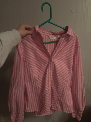 Rosa och vit randig skjorta - Säljer en rosa och vit randig skjorta med klassisk krage och långa ärmar. Skjortan har knappar framtill och är tillverkad i ett lätt bomullstyg som passar perfekt till en avslappnad stil. Storlek 44 men är som en M