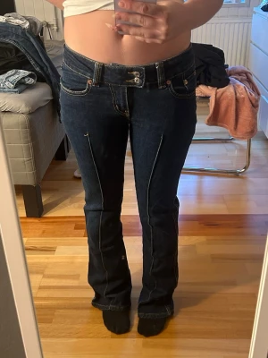 Mörkblå bootcut jeans med pressveck - Snygga mörkblå bootcut jeans med pressveck framtill och låg midja. Jeansen har klassiska fem fickor och kontrastsömmar. Perfekta för dig som gillar en retroinspirerad look och vill ha ett par jeans som sticker ut lite extra.