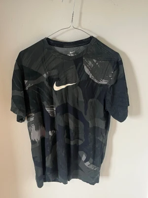 Svart camo t-shirt från Nike - Snygg svart t-shirt från Nike med camouflagemönster och vit logga på bröstet. Klassisk rund halsringning och korta ärmar. Perfekt för en sportig och avslappnad stil.