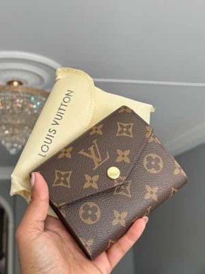 plånbok - Säljer en klassisk brun plånbok med ikoniskt monogrammönster i guld och brunt. Plånboken har kuvertformad flärp med guldfärgad knapp och är tillverkad i tåligt canvasmaterial. Perfekt för dig som gillar lyxiga accessoarer.