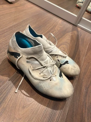 Vita fotbollsskor från Puma - Säljer ett par vita fotbollsskor från Puma med högt skaft och snörning. Skorna har svarta Puma-loggor på ovansidan och är tillverkade i syntetmaterial med mesh-liknande struktur. Perfekta för dig som vill ha stilrena och lätta skor på planen.