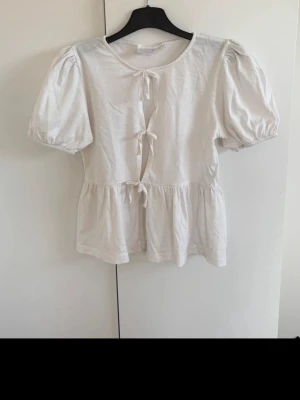 Vit blus med knyt och puffärm - Söt vit blus med puffärmar och tre knyt framtill. Blusen har en volang nedtill som ger en feminin passform. Perfekt för dig som gillar romantiska detaljer och enkel stil men uppklädd stil🤍 perfekt nu på sommaren. I storlek 158/164
