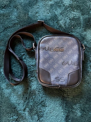 Mörkblå axelväska från Guess - Snygg mörkblå axelväska från Guess med justerbar axelrem. Väskan har ett mönster med Guess-loggor över hela ytan och detaljer i svart konstläder. Perfekt storlek för det viktigaste och stängs med dragkedja. Stilren och enkel att matcha till olika outfits.
