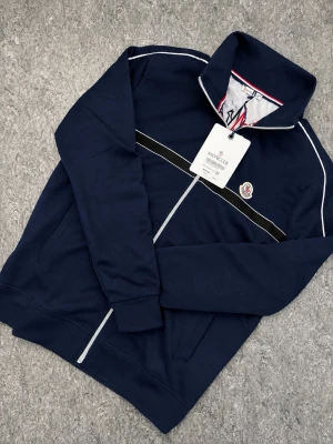 Moncler Ziptröja - Snygg marinblå tröja från Moncler med vit dragkedja, svarta och vita detaljer samt Moncler-logga på bröstet. Insidan har ett coolt bergsmotiv och stor Moncler-tryck. Tröjan har hög krage och ribbade muddar. Perfekt för dig som vill ha en stilren och exklusiv look.