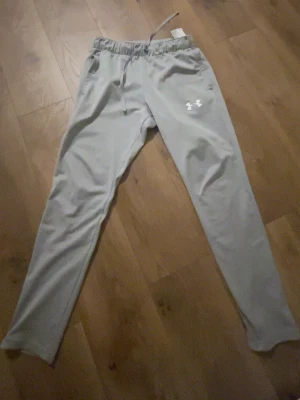 Grå mjukisbyxor från Under Armour - Snygga grå mjukisbyxor från Under Armour med smal passform och dragsko i midjan. Byxorna har ett vitt logotryck på låret och är tillverkade i ett mjukt och bekvämt material som passar perfekt för chill eller träning. Helt nya använd 2 gånger s men passar m