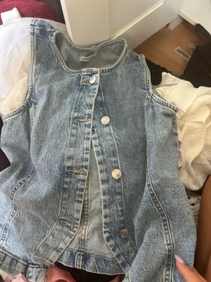 Ärmlös jeansväst i ljusblå denim - Säljer en snygg ärmlös jeansväst i ljusblå denim med fyra silverfärgade knappar framtill. Västen har klassisk rund halsringning och raka sömmar. Perfekt att styla över en t-shirt eller hoodie för en cool och avslappnad look.