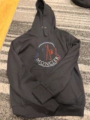 Svart Moncler hoodie - Snygg svart hoodie från Moncler med stor broderad logga i blått, vitt och rött på bröstet samt en mindre logga på ärmen. Hoodien har huva med dragsko och är tillverkad i ett strukturerat, mjukt material. Perfekt för dig som vill ha en exklusiv streetwear-look.