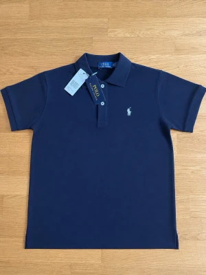 Mörkblå pikétröja från Polo Ralph Lauren - Klassisk mörkblå pikétröja från Polo Ralph Lauren med korta ärmar och vit broderad logga på bröstet. Tröjan har krage och knappar framtill. Perfekt till en avslappnad men ändå stilren look.
