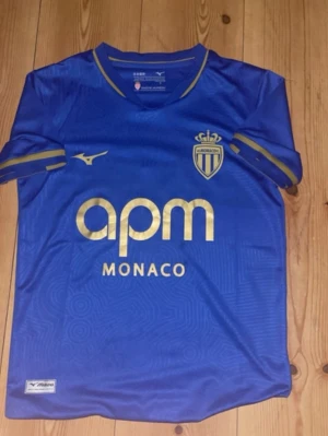 Fotboll tröja  - Snygg blå AS Monaco matchtröja från Mizuno med guldfärgade detaljer och klubbmärke på bröstet. Tröjan har rund hals, korta ärmar och tryckt sponsorlogga framtill. Tillverkad i lätt och ventilerande material, perfekt för fotboll eller träning.