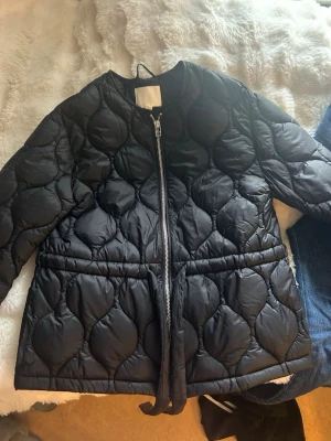 Svart quiltad pufferjacka med bälte - Säljer en svart pufferjacka med quiltat mönster och bälte i midjan. Jackan har rund halsringning, dragkedja framtill och en oversized look. Perfekt för kalla dagar och riktigt snygg till jeans eller kjol.knappt använd!