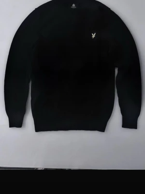 Lyle & Scott sweater - Klassisk svart stickad tröja med rund hals och en liten broderad logga i vitt på bröstet. Tröjan har långa ärmar och ribbade muddar vid ärmslut och nederkant. Perfekt basplagg för garderoben.