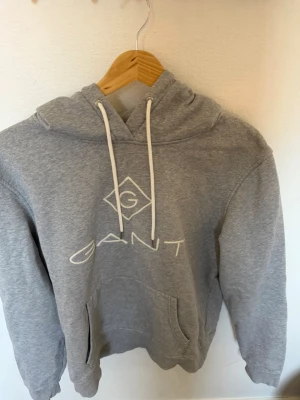 Grå hoodie från GANT med logga - Snygg grå hoodie från GANT med vit logga och dragsko i huvan. Klassisk känguruficka framtill och mjukt material som känns skönt mot huden. Perfekt för en avslappnad och trendig look.