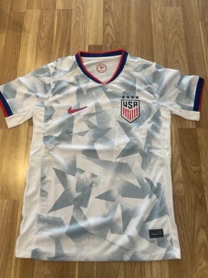 USA Nike fotbollströja vit/blå - Säljer en vit fotbollströja från Nike med USA:s landslagsemblem på bröstet. Tröjan har ljusblå stjärnmönster, röda och blå detaljer vid krage och ärmslut samt Nike-logga i rött. Perfekt för match eller träning!