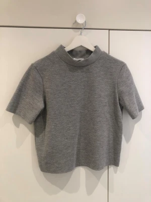 Grå polotopp med kort ärm - En stilren grå polotopp med korta ärmar och rak passform. Toppen har en hög krage och är tillverkad i ett mjukt, stickat material som ger en clean och modern look. Perfekt att matcha med jeans eller kjol för en enkel men snygg outfit.