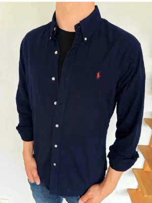 Mörkblå skjorta från Polo Ralph Lauren - Klassisk mörkblå skjorta från Polo Ralph Lauren med knappkrage och långa ärmar. Skjortan har vita knappar och den ikoniska röda logotypen broderad på bröstet. Tillverkad i ett mjukt bomullstyg som känns skönt mot huden.