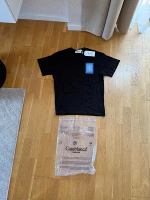 Svart t-shirt med blått tryck Casablanca - Snygg svart t-shirt från Casablanca med blått rektangulärt tryck på ryggen och mindre blå detalj på bröstfickan. Klassisk passform och rund hals. Perfekt för dig som gillar stilrena plagg med en twist.