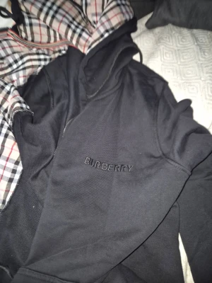 Svart hoodie från Burberry - Säljer en svart hoodie från Burberry med klassiskt rutigt foder i huvan och diskret broderad logga på bröstet. Hoodien har en avslappnad passform och är perfekt för dig som gillar stilrena plagg med exklusiv känsla.