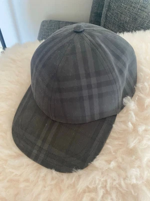 Burberry rutmönstrad keps - Snygg keps från Burberry i klassiskt rutmönster i mörkgrått/svart. Broderad 'BURBERRY' text baktill och justerbart spänne. Passar både dam och herr. Perfekt accessoar för en stilren look!