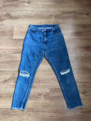 Blå slitna jeans med hål på knäna - Säljer ett par blå jeans med raka ben och slitningar på båda knäna. Jeansen har klassisk femficksmodell, hög midja och dragkedja med knapp. Perfekta för en avslappnad och trendig look.