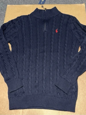 Mörkblå kabelstickad tröja Ralph Lauren - Mörkblå kabelstickad tröja från Ralph Lauren med halv dragkedja och hög krage. Tröjan har ett rött broderat logomärke på bröstet och ribbstickade muddar. Perfekt för dig som gillar klassisk och stilren look.