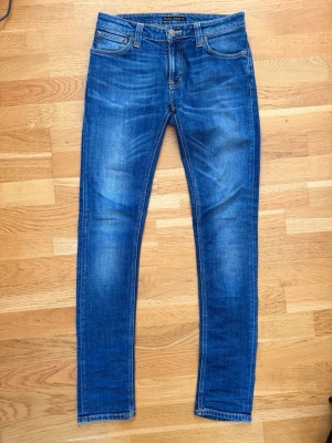 Blå Nudie jeans W29 L32 - Säljer ett par klassiska blå jeans från Nudie Jeans med smal passform och snygga slitningar framtill. Jeansen har fem fickor, tillverkade i mjuk denim med stretch för extra komfort. W29 L32. 
