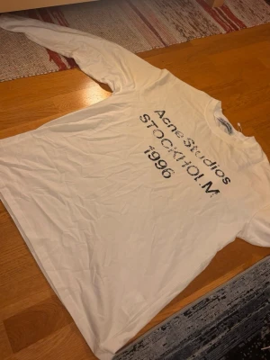 Vit långärmad t-shirt Acne Studios - Säljer en vit långärmad t-shirt från Acne Studios med svart tryck på bröstet där det står 'Acne Studios STOCKHOLM 1996'. Klassisk passform och rund hals. Perfekt för dig som gillar stilrena och trendiga plagg. Aldrig använd, står bara i garderoben.