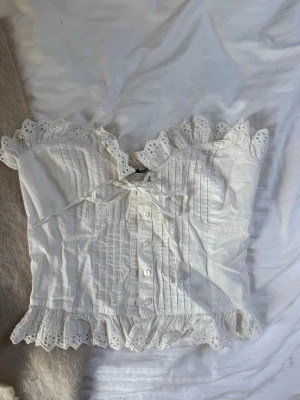 Vit korsettopp med spets och knyt - Supersöt vit korsettopp med spetsdetaljer och volangkant upptill och nedtill. Toppen har knappar framtill, dekorativ knytning vid bysten och är ärmlös. Perfekt för dig som gillar romantisk och feminin stil.