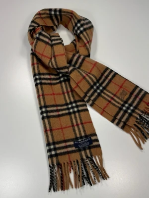 Beige rutig halsduk från Burberry - Klassisk beige halsduk från Burberry i ull med det ikoniska rutmönstret i svart, vitt och rött. Halsduken har fransar i ändarna och en vävd Burberry-etikett. Perfekt accessoar för att lyfta din outfit.0099