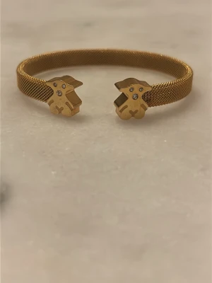 TOUS liknande guldigt armband med nallemotiv - Snyggt armband i guldtonad metall med meshstruktur och öppet avslut. Armbandet har två nalleformade detaljer i ändarna, dekorerade med små gnistrande stenar. Perfekt accessoar för dig som gillar lekfulla och trendiga smycken.