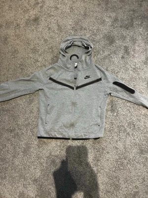 Grå Nike tech fleece  - Snygg grå hoodie från Nike med hel dragkedja och svart detalj över bröstet. Huva med hög krage och fickor på sidorna samt en extra ficka på ena ärmen. Perfekt för en sportig och avslappnad stil. Priset kan diskuteras.