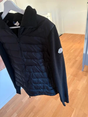 Svart quiltad jacka med stickade ärmar - Snygg svart jacka med quiltad front och stickade ärmar. Jackan har dragkedja framtill, mjuk krage och Moncler-logga på ärmen. Perfekt för dig som vill ha en stilren och bekväm jacka till vardags. Scann funkar