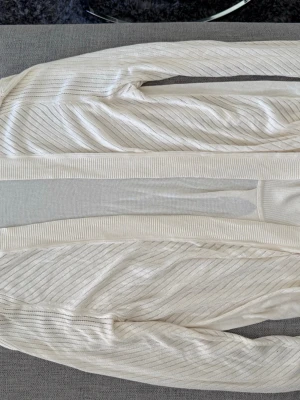 Vit lång kofta från H&M - Lång, öppen kofta från H&M i tunn, vit stickad kvalitet. Koftan har diskreta diagonala ränder och ribbade muddar vid ärmslut och nederkant. Perfekt att slänga över en topp för en chill look.