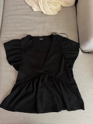 Svart v-ringad blus med volang - Snygg svart blus med djup v-ringning och volang nedtill. Toppen har korta puffärmar och är tillverkad i ett lätt, ribbat material som ger en trendig look. Perfekt att styla med jeans eller kjol för en cool outfit.