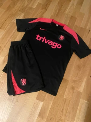 Chelsea fotbollsset - Säljer ett svart träningsset från Nike med Chelsea FC-logga och rosa detaljer. Setet består av t-shirt och shorts, båda med rosa tryck och logga samt trivago-tryck på bröstet. Perfekt för fotbollsträning eller gymmet.