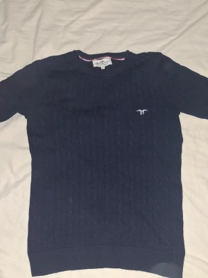 Mörkblå kabelstickad tröja Hampton Republic - Mörkblå kabelstickad tröja från Hampton Republic med rund halsringning och diskret logga broderad på bröstet. Tröjan har ribbade muddar och klassisk passform. Perfekt för dig som gillar stilrena och tidlösa plagg.