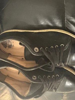Svarta låga sneakers med vita sulor - Säljer ett par svarta sneakers med lågt skaft och vita sulor. Skorna har svarta snören, metallfärgade öljetter och en blank tådel. Insidan är beige och materialet ser ut att vara en mix av textil och syntet. Perfekta för en clean och enkel stil.
