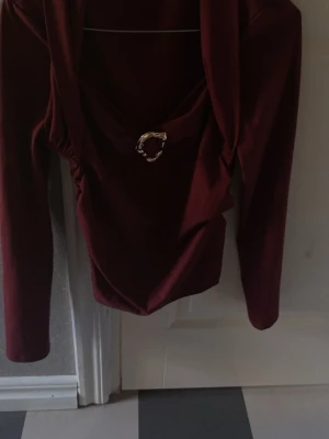 Vinröd långärmad topp med detalj - Snygg vinröd topp med långa ärmar och en dekorativ guldfärgad ring framtill vid urringningen. Toppen har en draperad effekt och är perfekt för dig som vill sticka ut lite extra.