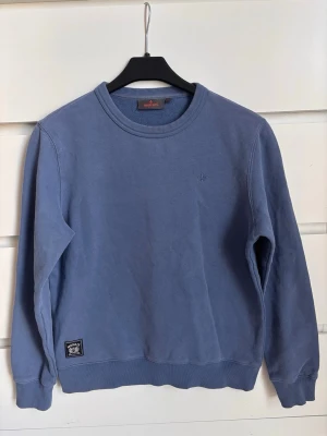 Blå sweatshirt från Morris Stockholm  - En stilren blå sweatshirt från Morris Stockholm med rund hals och ribbade muddar vid ärmslut och nederkant. Tröjan har en liten diskret logga på bröstet och en patch nertill. Perfekt för en avslappnad look och enkel att matcha.