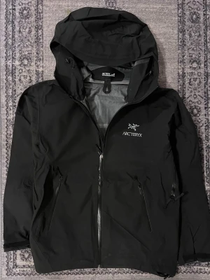 Arc'teryx - Säljer en svart regnjacka från Arc'teryx med huva och dragkedja framtill. Jackan har två snedställda fickor med dragkedja och logga broderad på bröstet. Materialet är tunt och vindtätt, perfekt för blåsiga dagar.
