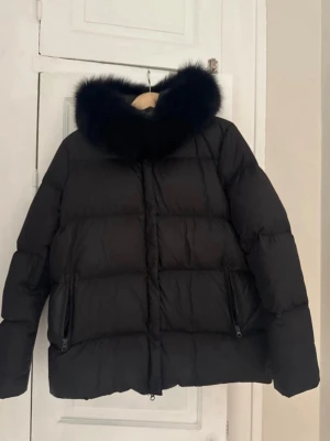Svart dunjacka med pälskantad huva meotine - Snygg svart dunjacka med stor, fluffig huva kantad med svart päls. Jackan har två dragkedjeförsedda fickor framtill och quiltad design som ger en puffig look. Perfekt för kalla vinterdagar och riktigt varm. Storlek s/m passar xs-m.