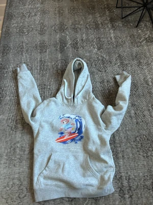 Grå hoodie med polo-tryck - Säljer en grå hoodie med stor huva och magficka. På bröstet finns ett färgglatt Björn tryck med en surfande karaktär och blå vågor. Hoodien har en avslappnad passform och är gjord i mjukt bomullsmaterial. Perfekt för dig som gillar streetwear och old money.