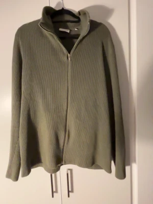 Olivgrön ziptröja Lindbergh - Säljer en olivgrön ribbstickad ziptröja från Lindbergh i relaxed fit. Tröjan har hög krage, dragkedja framtill och långa ärmar. Perfekt för dig som gillar en enkel men snygg stil. Passar till det mesta och är superbekväm. Lite små slitage men enkelt att laga med att sy
