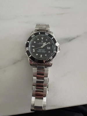 Silverfärgad klocka med svart urtavla - Stilren klocka i silverfärgat metallarmband och svart urtavla med vita detaljer. Klockan har en klassisk rund form och tydliga markeringar på boetten. Perfekt accessoar för dig som gillar en tidlös look.