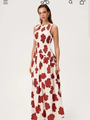 Adoore klänning - Långklänning i vit bas med stora röda blommor. Klänningen har ärmlös design och rund halsringning. Materialet ser lätt och luftigt ut, perfekt för varma dagar. Passformen är normal och klänningen har en romantisk och somrig känsla.