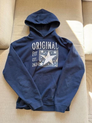 Scandi Original archive hoodie - Snygg mörkblå hoodie med stor vit stjärna och texten 'ORIGINAL FIT EST. 2024' framtill. Hoodien har huva och en klassisk magficka. Perfekt för dig som gillar en avslappnad och cool stil. Materialet känns mjukt och bekvämt. Obs saknar snören. Hoodien säljs inte längre så den är sällsynt