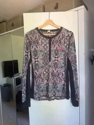 Mönstrad långärmad tröja med knappar - Snygg långärmad tröja med färgglatt paisleymönster i svart, rosa, turkos och vitt. Tröjan har rund halsringning och knappslå framtill samt svarta paneler längs ärmarna. Perfekt för dig som gillar unika mönster och bekväma plagg. Från märket Odd Molly i storlek 3 som enligt deras storlekguide motsvarar en storlek L. Mycket fint skick, jag hittar inga synliga anmärkningar eller defekter. 